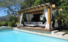 a gazebo next to a swimming pool with lounge chairs and curtains on the side