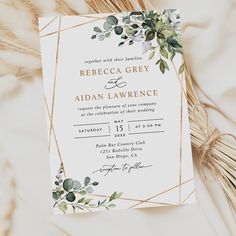 an elegant wedding card with greenery and gold foil on the front, sitting next to some dried grass