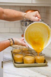 a woman pours orange juice into small glass jars on a tray in the kitchen