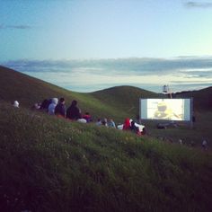 people sitting in the grass watching an outdoor movie on a hill with a projector screen