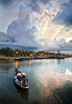 two people in a small boat on the water under a cloudy sky with palm trees