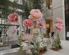 pink flowers are in vases on the ground outside an office building with people walking by