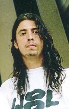 a man with long hair wearing a white t - shirt and looking at the camera