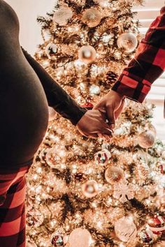 a pregnant woman and man holding hands in front of a christmas tree