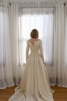 a woman standing in front of a window wearing a long white dress and looking out the window