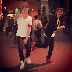 two young men standing next to each other in front of a building with people walking around