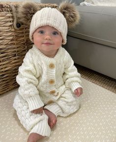 a baby sitting on the floor wearing a white sweater and hat with fur pom - poms