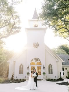 a bride and groom standing in front of a white church with lights on the windows