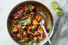 a pan filled with meatballs and onions on top of a marble table next to lime wedges