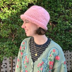 a woman wearing a pink knitted hat standing in front of some bushes and trees