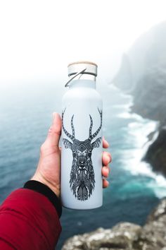 a person is holding a water bottle with a deer head on it and the ocean in the background