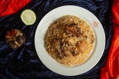 a white plate topped with rice and meat next to a slice of lime on a blue cloth