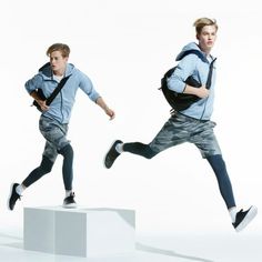 two young men in blue shirts and grey shorts are jumping on white pedestals, one is carrying a black backpack