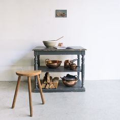 an old table with bowls on it and a stool next to it in front of a white wall