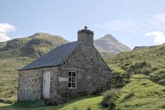 an old stone building on the side of a grassy hill with mountains in the background