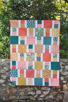 a patchwork quilt hanging on a stone wall with trees in the backgroud