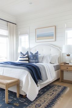 a white bed with blue and white pillows on top of it in a bedroom next to two windows