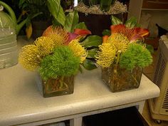 two clear vases with yellow and green flowers on a table next to other plants