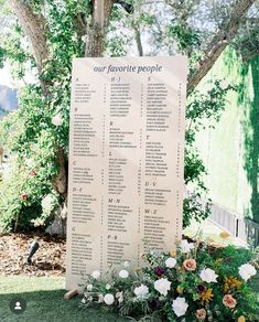 a sign that is sitting in the grass with flowers on it and some trees behind it