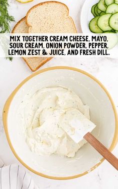 a bowl filled with cream cheese next to sliced cucumbers and bread on a table