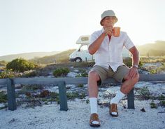 a man sitting on a bench with a cup of coffee in his hand and a camper behind him