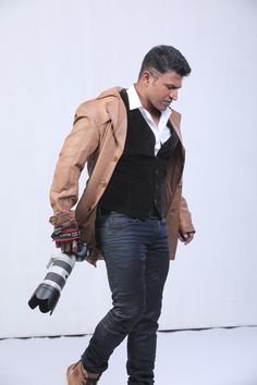 a man in black shirt and brown jacket holding an umbrella while standing next to a white wall