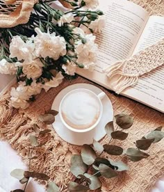 a cup of coffee on top of a saucer next to an open book and flowers