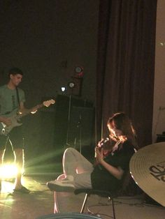 two people are playing instruments in a dark room with lights coming from behind them and one person is sitting on a chair