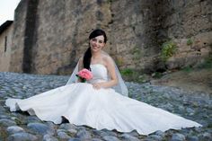 a woman in a wedding dress is sitting on the cobblestone road holding a flower