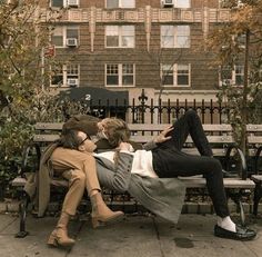 two people sitting on a bench in front of a building and one person laying down