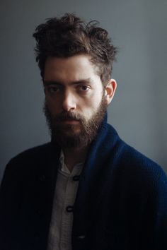 a man with a beard wearing a cardigan and looking at the camera while standing in front of a gray wall