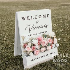 a welcome sign with pink and white flowers on it sitting in the grass next to a field