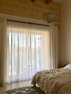 a bed sitting next to a window covered in white sheer curtains and a light fixture