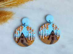 a pair of earrings with trees and houses painted on them sitting next to dried grass