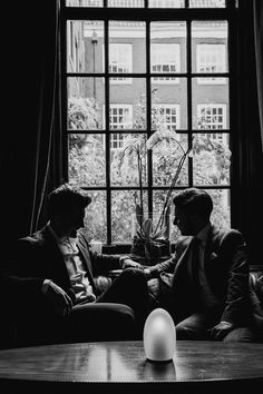 two men sitting on a couch in front of a large window talking to each other