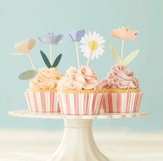 three cupcakes on a cake stand decorated with paper flowers and daisols