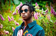 a man with dreadlocks and sunglasses standing in front of some purple flower bushes