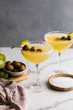 two martinis with olives and salt in them on a marble countertop next to limes