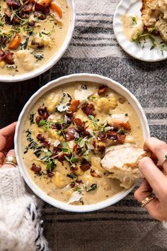 two hands holding a bowl of soup with bread on the side and other dishes in the background