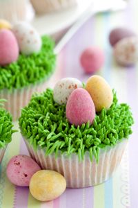 three cupcakes decorated with grass and eggs on a striped tablecloth next to other desserts