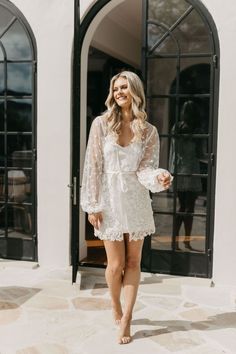 a woman standing in front of a door wearing a white lace robe with sheer sleeves