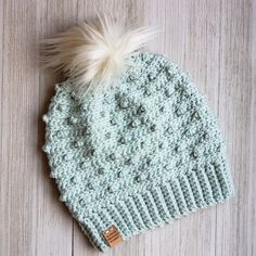 a crocheted hat with a white pom - pom sits on a wooden surface