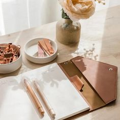 an open notebook and pen on a table next to a vase with flowers in it