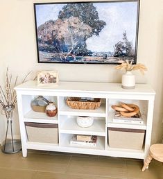 a white shelf with baskets and pictures on it in front of a wall mounted painting