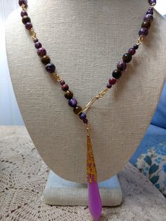 A lariat necklace made of purple jasper and tiger's eye beads on a gold-plated chain, with a purple and white resin pendant in a gold-plated iron setting. The necklace is 25 3/4 inches long from end to end and the pendant glows blue in the dark if exposed to UV light. To where it, the pendant goes through the gold-plated loop on the opposite end, and it can be tightened to the wearer's liking. Gold Wire Wrapped Lariat Necklace As Gift, Gold Teardrop Agate Necklace, Bohemian Gold Lariat Necklace With Gemstone Beads, Gold Bohemian Lariat Necklace With Gemstone Beads, Purple Wire Wrapped Long Drop Jewelry, Handmade Purple Lariat Necklace, Adjustable Gold Lariat Necklace With Natural Stones, Unique Lariat Necklace With Natural Stones For Gifts, Spiritual Gemstone Beaded Lariat Necklace As Gift