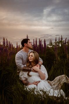 a man and woman sitting in tall grass with purple flowers on the ground behind them