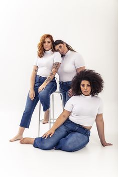 three women in white shirts and jeans sitting on a stool with their arms around each other