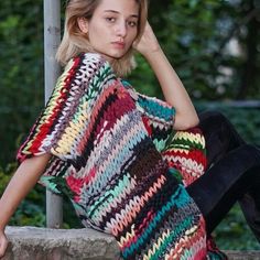 a woman sitting on a stone wall wearing a multicolored crochet sweater