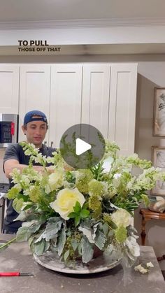 a man standing in front of a vase filled with white and green flowers on top of a kitchen counter