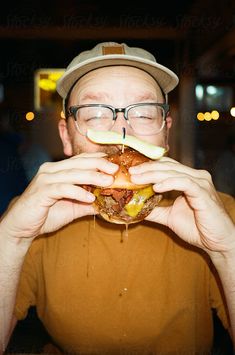 a man with glasses and a hat is biting into a large hamburger while holding it up to his mouth